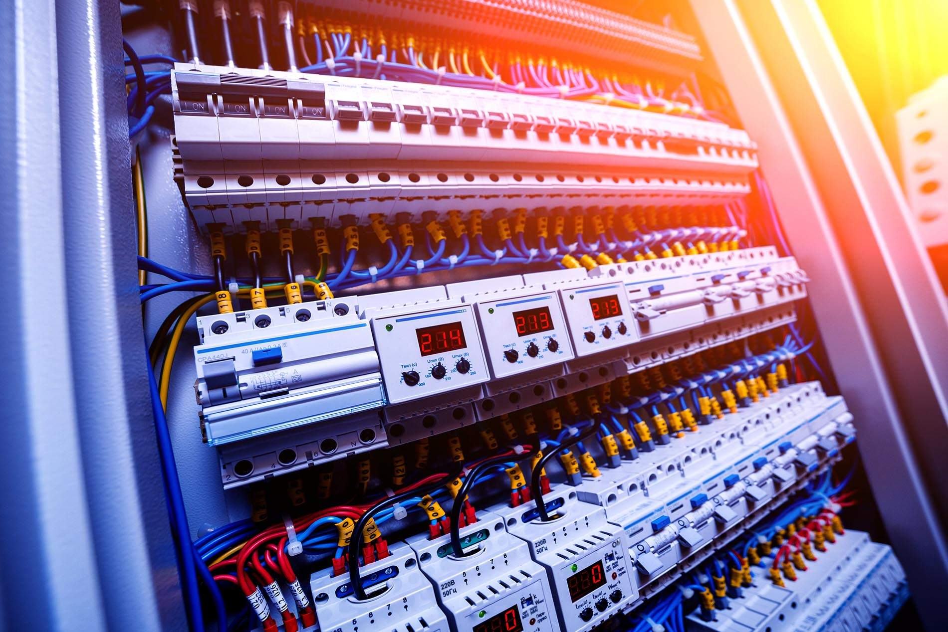 Rows of circuit breakers, digital meters and wiring inside an open distribution board, lit in orange and blue