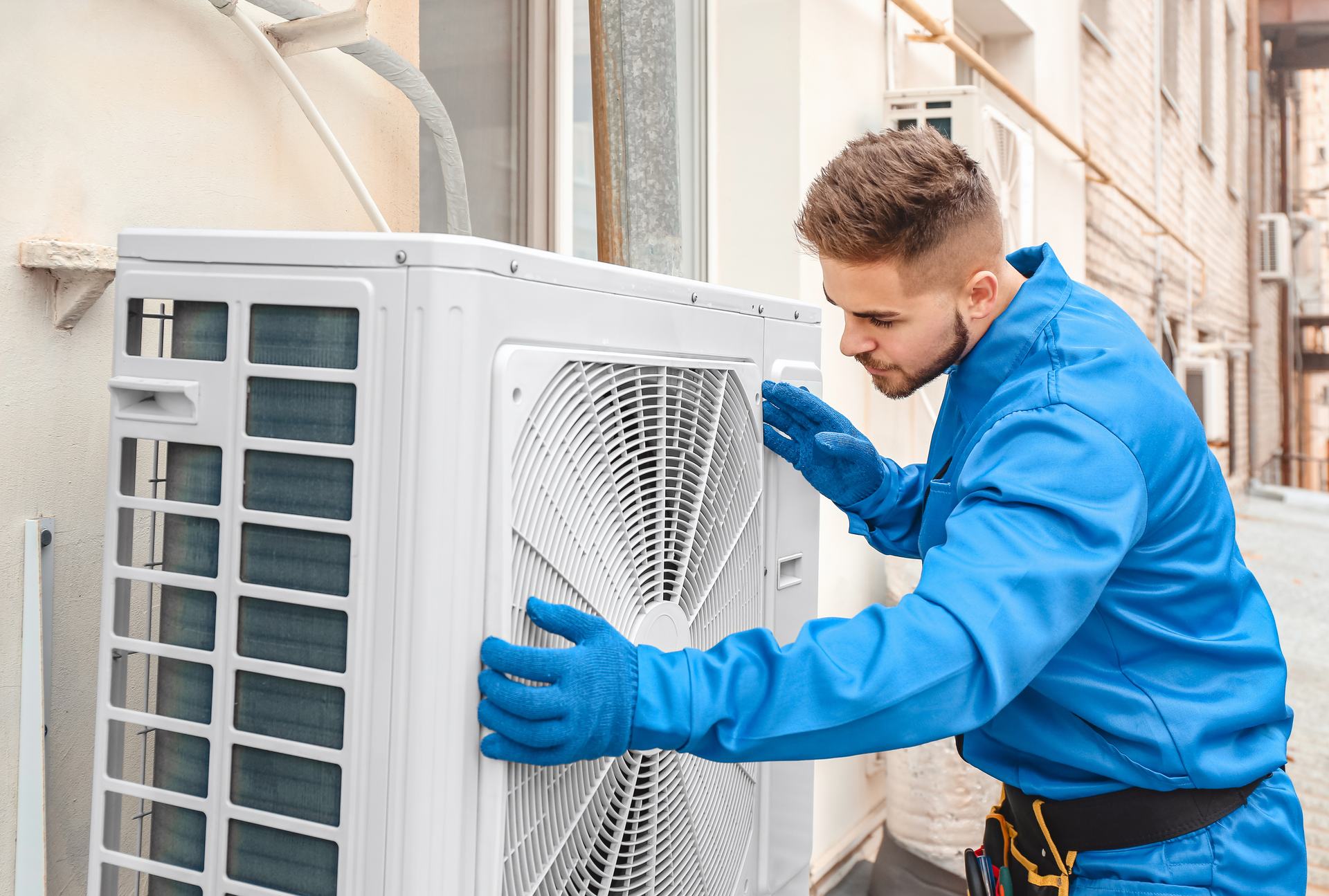 Technician in blue overalls and gloves working on a heat pump outdoor unit beside a house