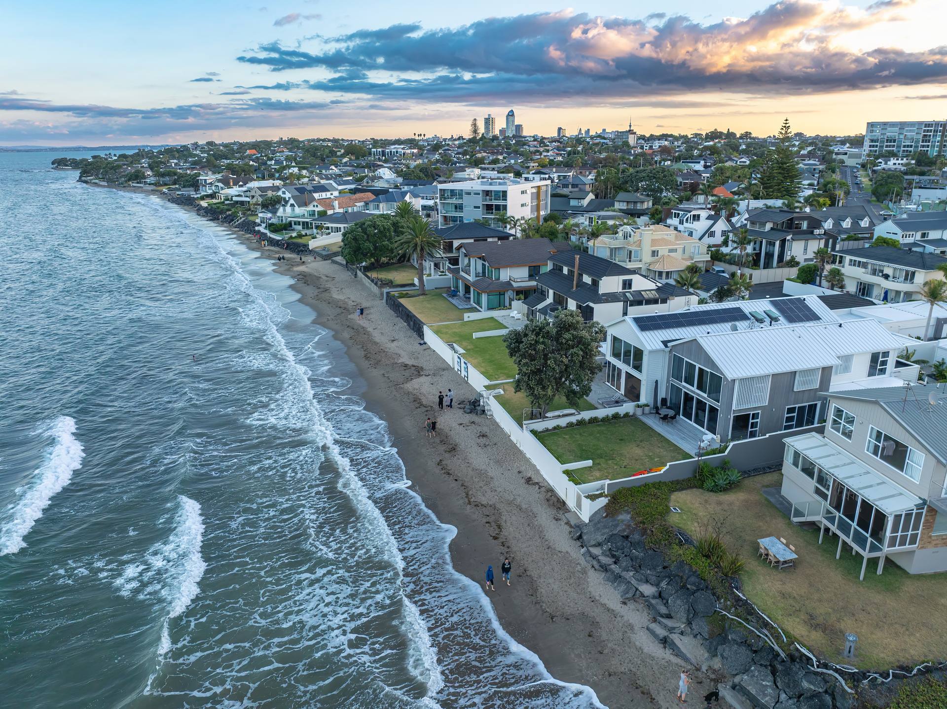 Aerial view at dusk along a sandy beach lined with houses, lawns and a rock seawall, with apartment towers on the skyline