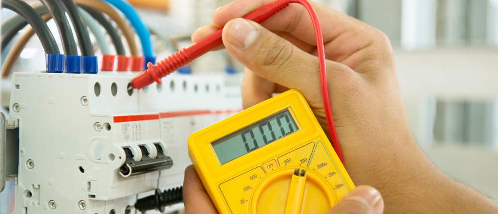 A hand holding a yellow digital multimeter while its red probe touches a circuit breaker on a DIN rail
